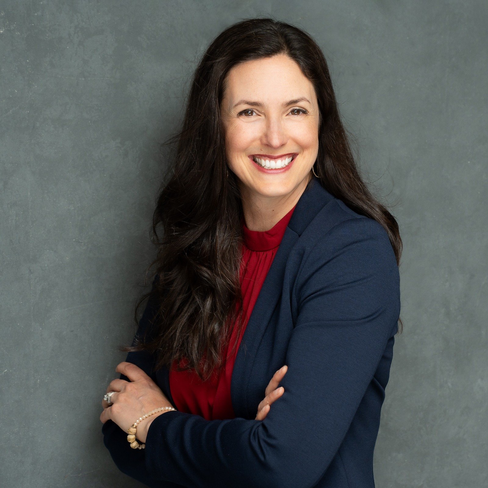Smiling professional woman in a navy blazer with a red blouse, arms crossed against a gray backdrop.