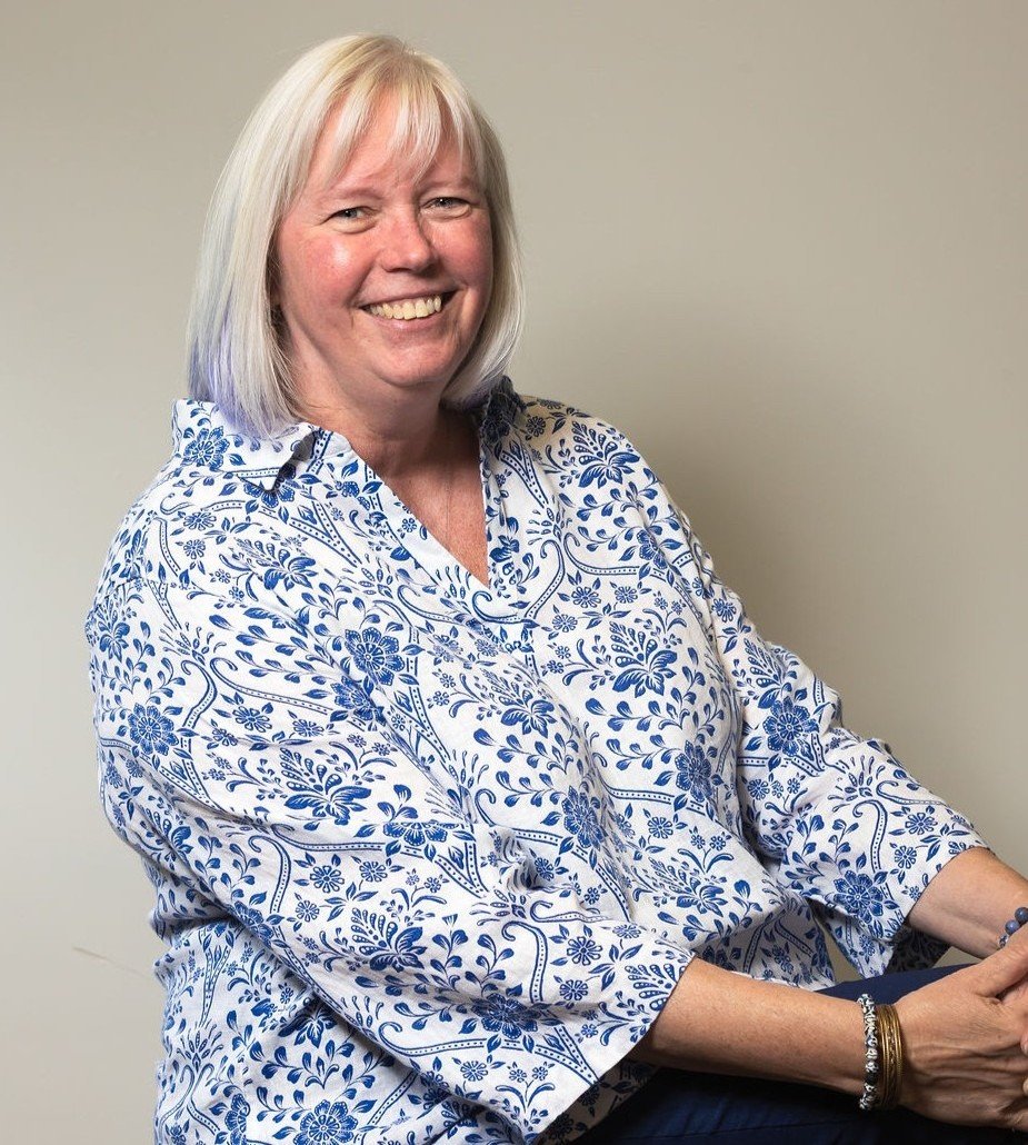 Smiling woman with shoulder-length light hair wearing a white and blue floral blouse, seated indoors.