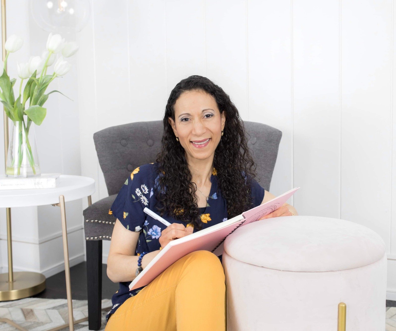 Woman with long curly hair sits on a pale pink ottoman, writing in a pink notebook and smiling at the camera in a bright, white-room setting.